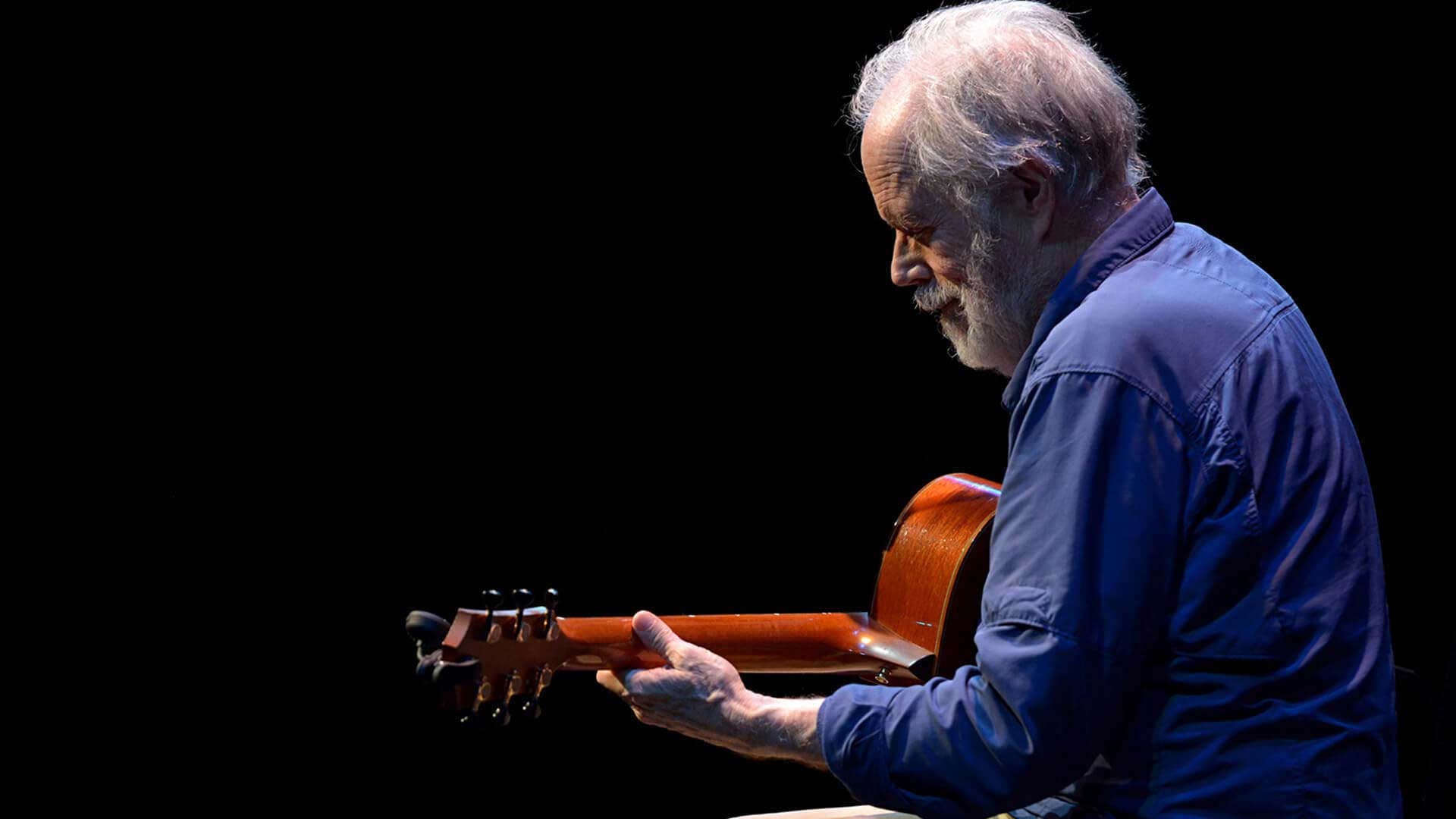 Leo Kottke beneath a spotlight on an darkened stage, working his stunning fingerpicking style on an acoustic guitar.